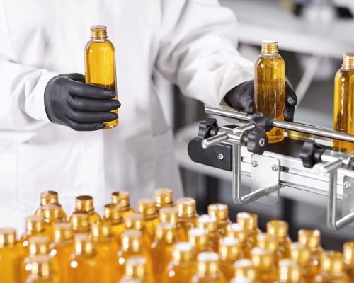 Photo of production new kind of cosmetics by professional researcher. Young scientist standing isolated over factory background dressed in gown and black gloves holding bottles with yellow liquid