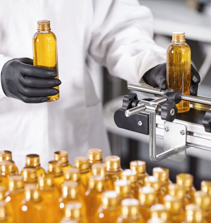 Photo of production new kind of cosmetics by professional researcher. Young scientist standing isolated over factory background dressed in gown and black gloves holding bottles with yellow liquid